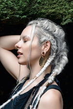 A woman with platinum blonde hair styled in intricate braids poses with her eyes closed and hand behind her head. She wears large, ornate earrings and an AYA Septum Clicker Shipibo Pattern by Kaya Movement. The background is dark with a hint of greenery at the top.