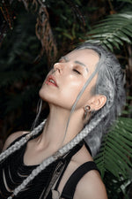 A person with long, grey braided hair and dark eye makeup is standing outdoors against a backdrop of green foliage. They are adorned with multiple earrings, including the Kaya Movement KAMO Biomech Teardrop Clicker Septum Ring in Silver (16 gauge) and stud earrings, while their eyes are closed and their head is tilted slightly back.