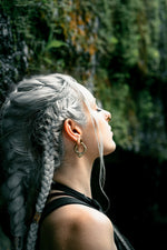 A woman with long, braided, silver hair and gold MARVDASHT Small Cyborg Hoop Clicker Ear Weights in 2 gauge by Kaya Movement stands in profile against a backdrop of lush greenery and rocks. She is looking upward with a calm expression, dressed in a black top, and the natural light highlights her hair and face.