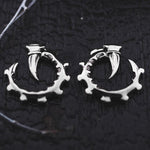 The GIGER Biomechanical Open Ended Hoop Ear Weights in Silver 925 from Kaya Movement are designed to resemble curved claws gripping an intricate, spiked ring. The jewelry rests on a dark, textured surface with hints of white threads at the edges, accentuating the contrast between the metal and the background.