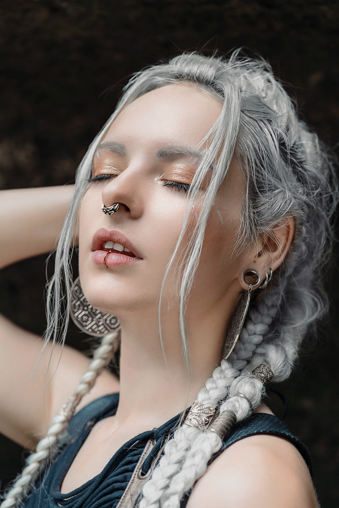 A person with long, braided white hair stands with eyes closed and one arm raised. They are wearing intricate sterling silver 925 earrings, an AYA Septum Clicker Shipibo Pattern Inspired by Silver 925 from Kaya Movement (16 gauge), and a lip piercing. The expression on their face appears serene. A dark and blurred background complements their appearance.
