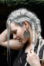 A person with long, platinum braided hair adorned with silver bands is shown in profile. They wear large, intricate circular earrings made from Sterling Silver 925 and have multiple facial piercings, including an AYA Septum Clicker Shipibo Pattern by Kaya Movement. Their hand gently touches their hair as they are dressed in black clothing.