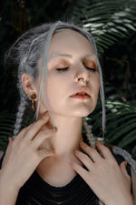 A person with long, braided gray hair is posed against a backdrop of green leaves. They have multiple piercings, including Kaya Movement’s KAMO Biomech Teardrop Septum Ring in Gold (16 gauge), a nose ring, and a lip ring. Their makeup includes shimmering eyeshadow. They are gently touching their neck with both hands.