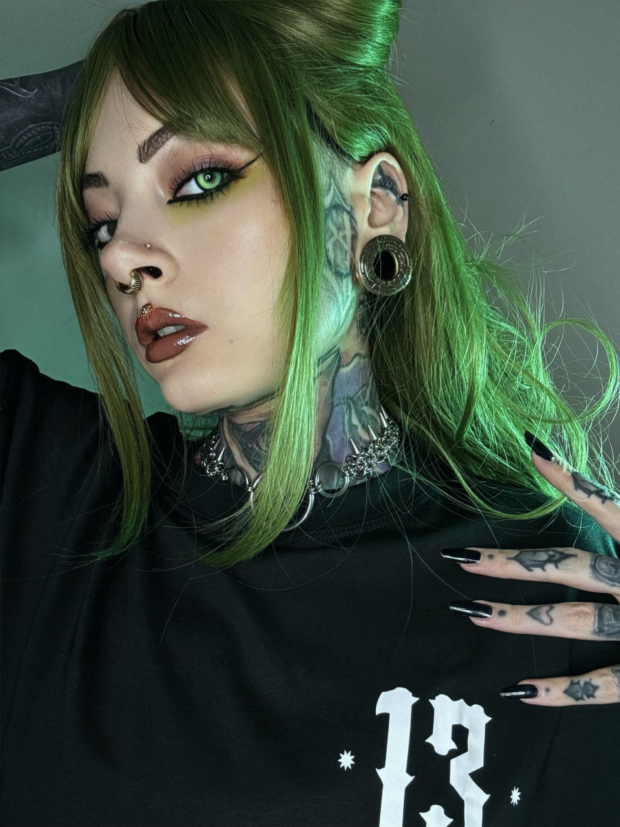 A person with green hair, a nose ring, and a large gold tunnel in their stretched ear. They have tattoos on their neck and hands. Their look combines bold, futuristic, and cyberpunk elements, with sharp, detailed styling