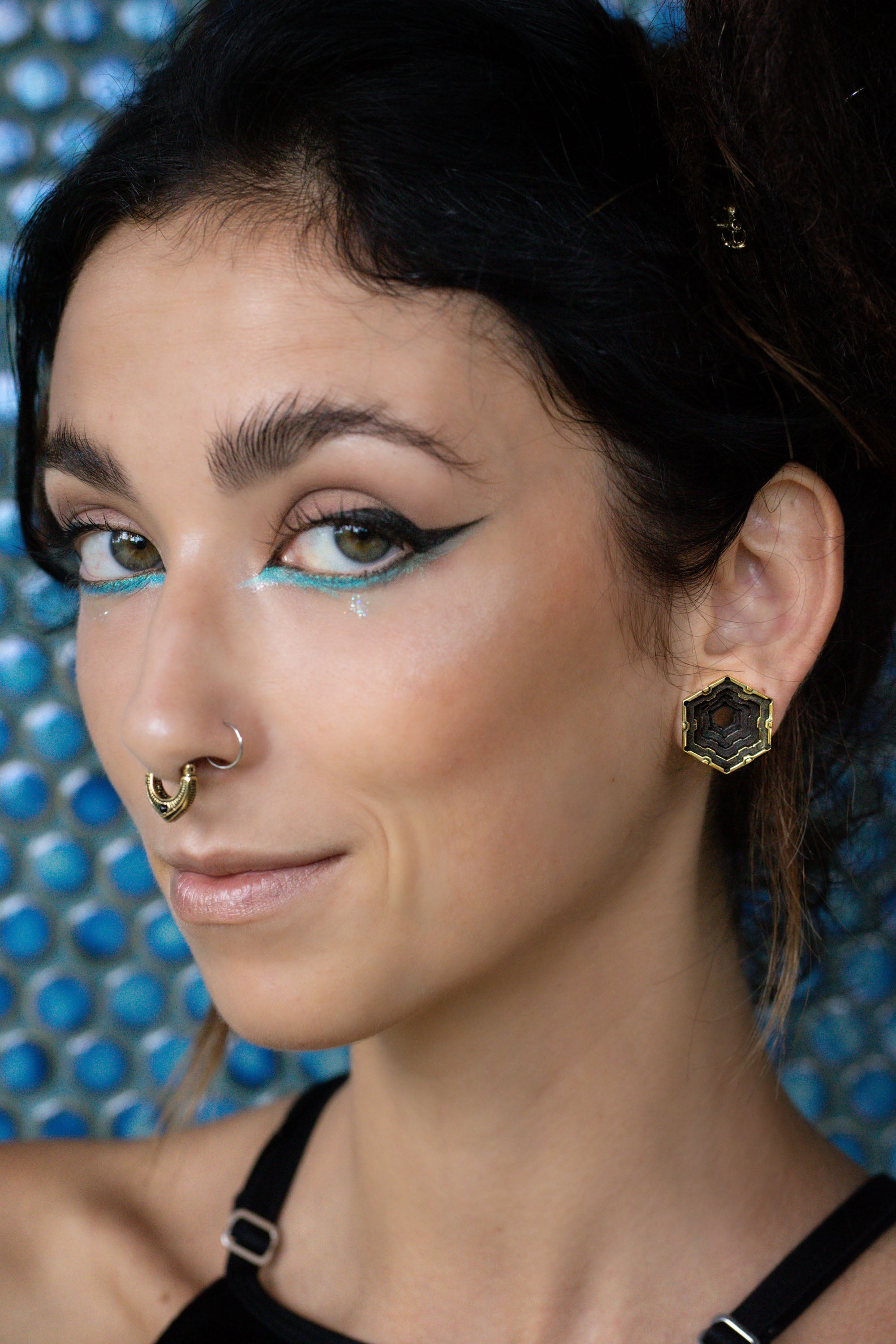 Woman with turquoise eyeliner and dramatic black cat-eye makeup, smiling subtly. She wears a gold septum ring and hexagonal earrings with a black geometric design, set against a textured blue background.