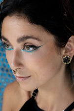 Close-up of a woman with bold turquoise and black winged eyeliner, wearing a gold septum ring and hexagonal black-and-gold earrings. Her expression is serene, and her brown eyes are softly gazing to the side.
