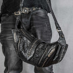 A person wearing distressed black jeans and a black belt with silver studs is holding the Nebula Unisex Leather Off Shoulder Cross Body HandBag from Kaya Movement, adorned with zippers, studs, and intricate silver detailing. The upper body is partially visible, focusing on the midsection and the bag, reminiscent of attire seen at dystopian festivals.