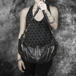 A person dressed in a sleeveless top with a black hexagonal pattern and dark pants holds the Kaya Movement Nebula Unisex Leather Off Shoulder Cross Body HandBag in front of a textured, dark gray backdrop. The top of the person's head is not visible in the image. Multiple bracelets adorn both wrists, reminiscent of accessories at dystopian festivals.