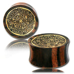 Two MAYA Round Saddle Plugs in Gold & Wood by Kaya Movement, featuring intricate golden geometric designs on the front. The wooden gauges are rounded with a slightly concave shape and a smooth, dark brown finish. The reflective surface beneath them shows a faint mirror image of the plugs. Available in sizes from 12mm to 25mm gauge.