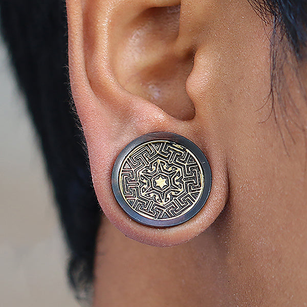 A close-up of a person's ear wearing Kaya Movement's MAYA Round Saddle Plugs in Gold & Wood. The round plug features an intricate, geometric mandala design in gold and black colors. The person's hair is short, and the background is blurred.