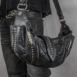 A person is carrying the Nebula Unisex Leather Off Shoulder Cross Body HandBag by Kaya Movement, a custom black leather bag adorned with metal studs and textured patches, perfect for dystopian festivals. The Nebula features multiple intricate details, like a ribbed design and decorative stitching. The person complements their look with black clothing and a studded belt.