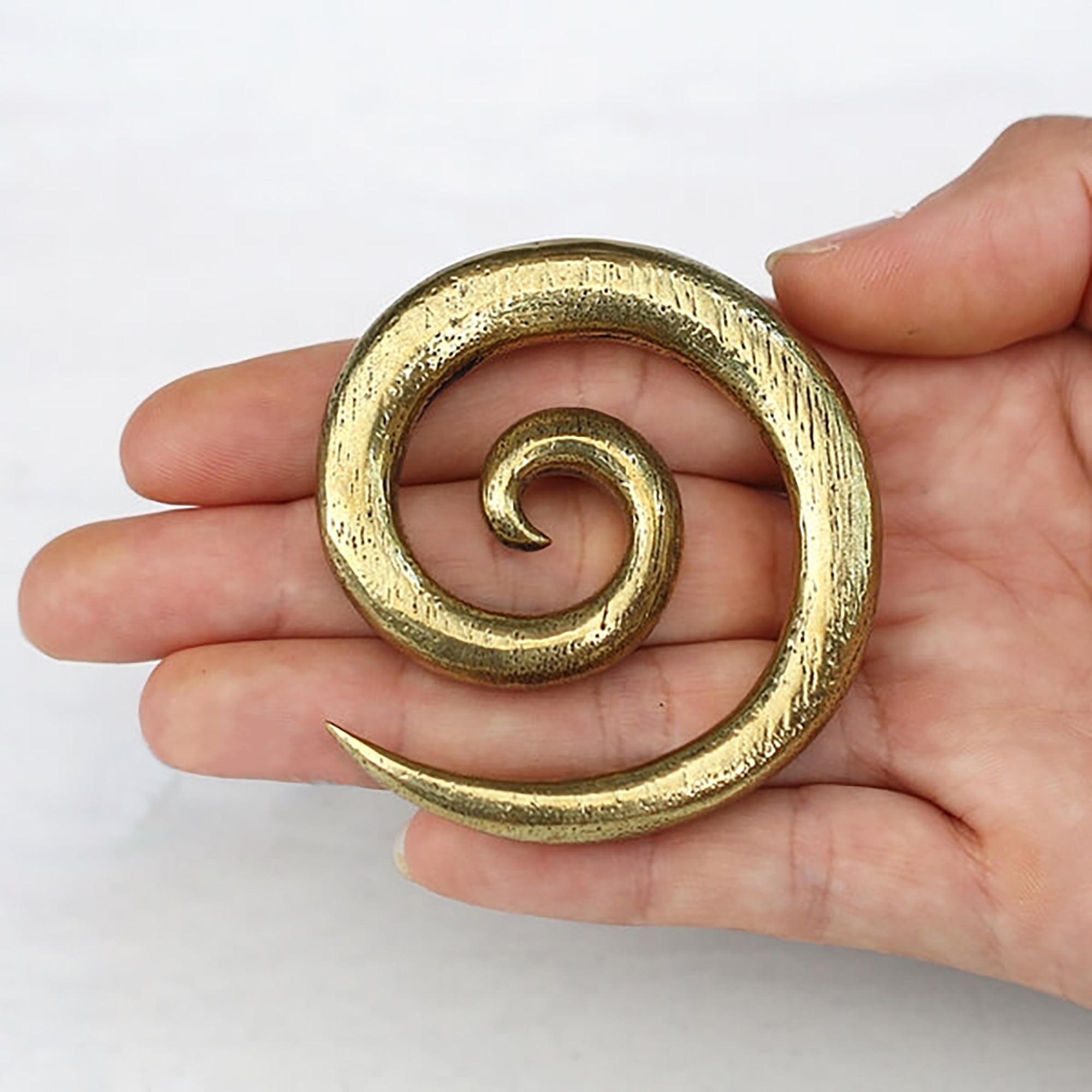 A hand holds the SPIRAL Minimalist Ear Weights in Gold by Kaya Movement against a plain background. These 00 gauge ear weights, reminiscent of brass gauge design, form a smooth spiral tapering to a point at one end. The piece has a rough, textured surface that adds an artisanal touch.
