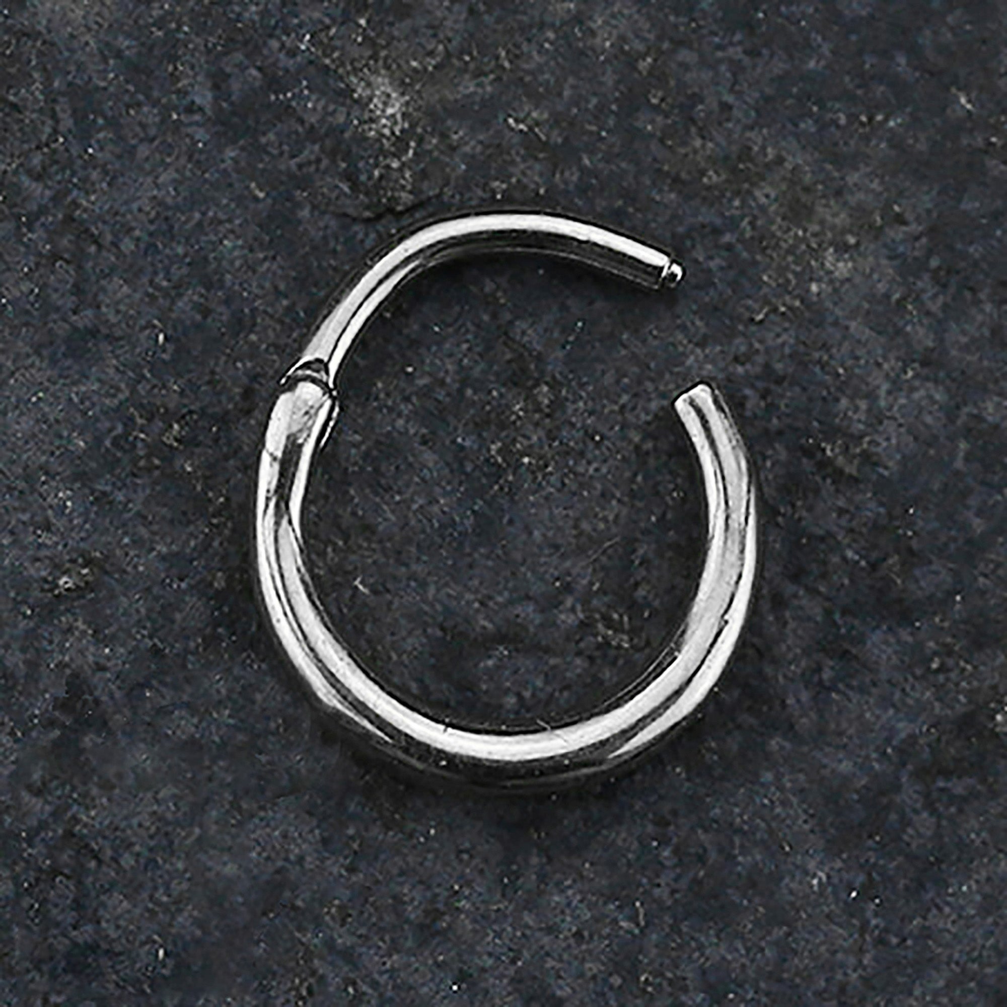 Lying on a dark, textured surface is a small AMAO Clicker Nose Ring by Kaya Movement, made from 316L surgical steel with a smooth silver finish. The 16 gauge ring is open at one end, indicating it's an unworn piece of versatile body jewelry.