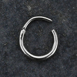 Lying on a dark, textured surface is a small AMAO Clicker Nose Ring by Kaya Movement, made from 316L surgical steel with a smooth silver finish. The 16 gauge ring is open at one end, indicating it's an unworn piece of versatile body jewelry.