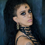 A person with long, dark, curly hair and dramatic makeup featuring black lines, dots, and a crescent moon on their face. They have a septum piercing and are wearing the TECHRE Futuristic Sci Fi Hexagon Keyhole Ear Weights in Gold (0 gauge) by Kaya Movement along with a black strappy top or harness. The background is dark and blurred.