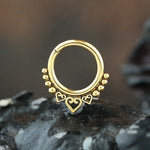 The ALYA Lotus Flower Clicker Septum Ring in Gold by Kaya Movement, a 16-gauge piece of body jewelry featuring a gold brass body adorned with small golden beads along the top and three heart-shaped charms hanging at the bottom, is displayed against a dark, textured background. The design is intricate and elegant.