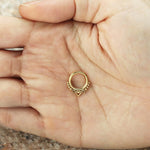 A close-up of a hand holding the ALYA Lotus Flower Clicker Septum Ring in Gold by Kaya Movement. This 16-gauge ring features three dangling heart charms evenly spaced around the band, with intricate lotus designs etched into its surface. The hand is open, palm facing upwards to showcase the details against a softly blurred background.