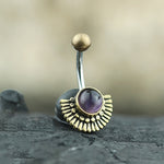 A close-up image of the OSIRIS Marquise Belly Piercing Ring in Gold & Amethyst by Kaya Movement, showcasing a gold, textured wing-shaped design. At the center is a round amethyst encased in a gold setting. The curved barbell, made of surgical steel and designed for 14 gauge piercings, is topped with a small golden sphere.