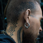 A close-up side profile of a person with a partially shaved head and intricate, zigzag tattoos on the scalp and neck. The person has a beard and is wearing Kaya Movement's MARVDASHT Small Biomechanical Clicker Ear Weights in 925 Silver, designed in a 2 gauge size that add subtle sci-fi vibes. The background is blurred and dark.