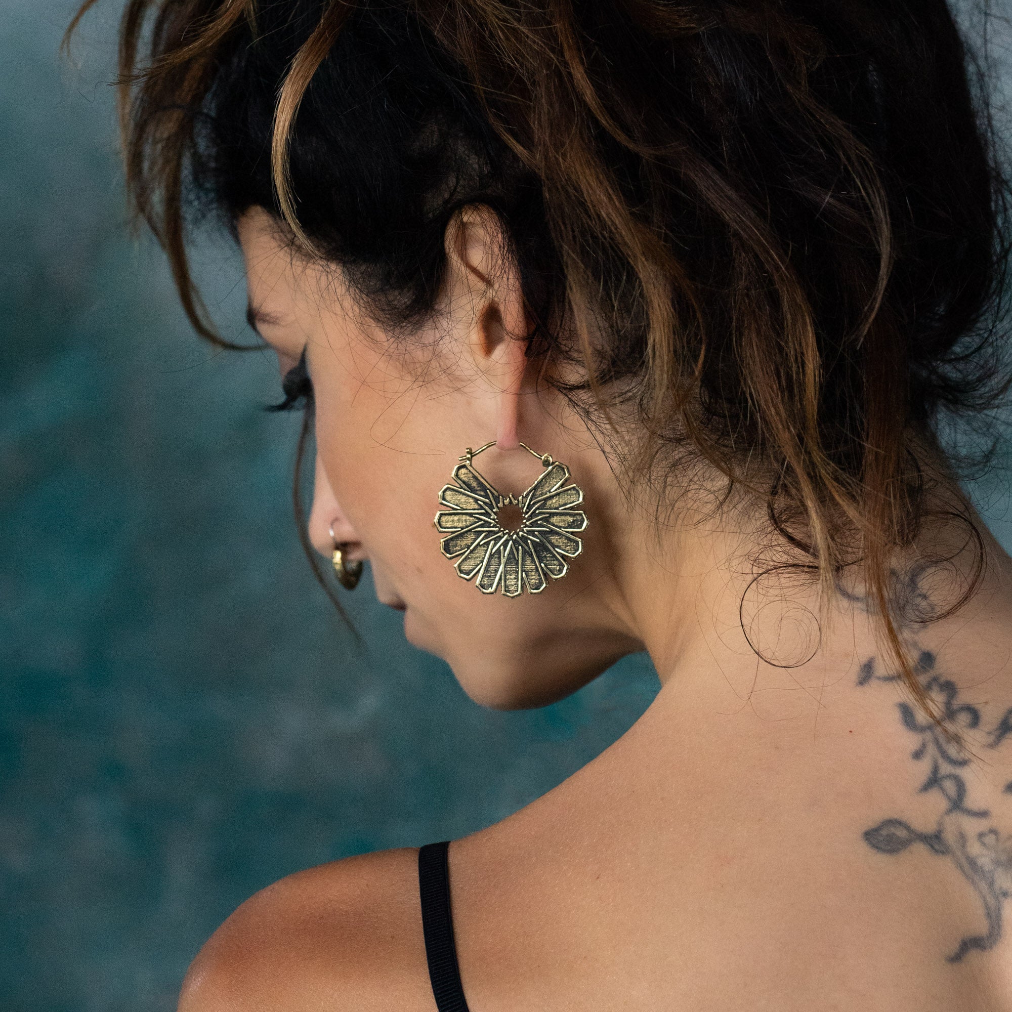A woman with dark hair pulled up loosely, showing her back and side profile, wears the striking MOSAIC Ethnic Mandala Disc Hoop Earrings in Gold from Kaya Movement. The large, intricate boho statement earrings shaped like flowers complement her nose ring beautifully. A floral tattoo is visible on her left shoulder against a backdrop of blurred blue and green tones.