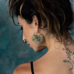 A woman with dark hair pulled up loosely, showing her back and side profile, wears the striking MOSAIC Ethnic Mandala Disc Hoop Earrings in Gold from Kaya Movement. The large, intricate boho statement earrings shaped like flowers complement her nose ring beautifully. A floral tattoo is visible on her left shoulder against a backdrop of blurred blue and green tones.