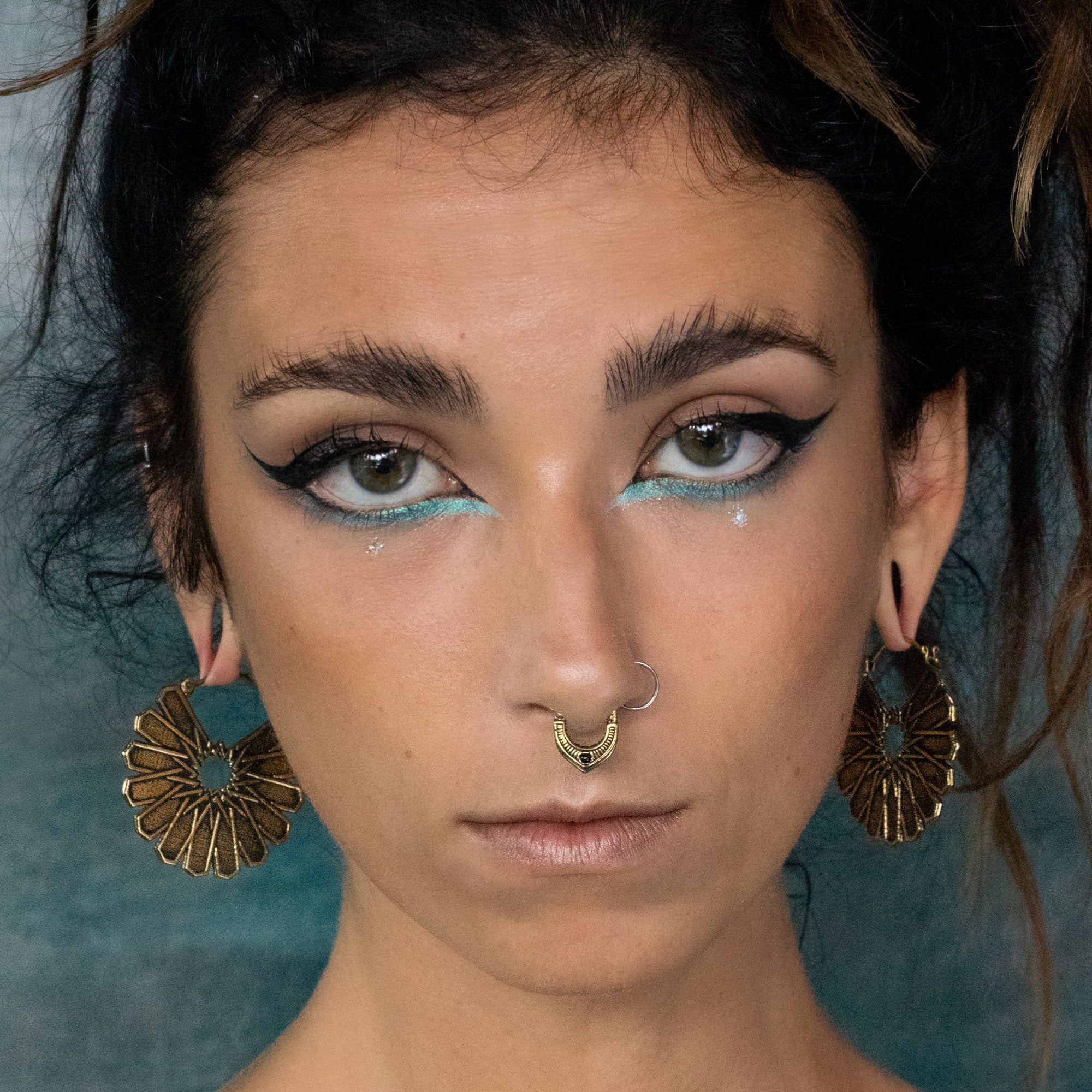 Close-up of a person with striking makeup, featuring bold black eyeliner and light blue eyeshadow. They have a nose ring, stretched earlobes adorned with Kaya Movement's MOSAIC Ethnic Mandala Disc Hoop Earrings in Gold (16 gauge), and a serious expression. The background is a textured teal color.
