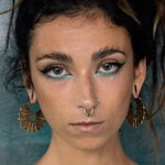 Close-up of a person with striking makeup, featuring bold black eyeliner and light blue eyeshadow. They have a nose ring, stretched earlobes adorned with Kaya Movement's MOSAIC Ethnic Mandala Disc Hoop Earrings in Gold (16 gauge), and a serious expression. The background is a textured teal color.