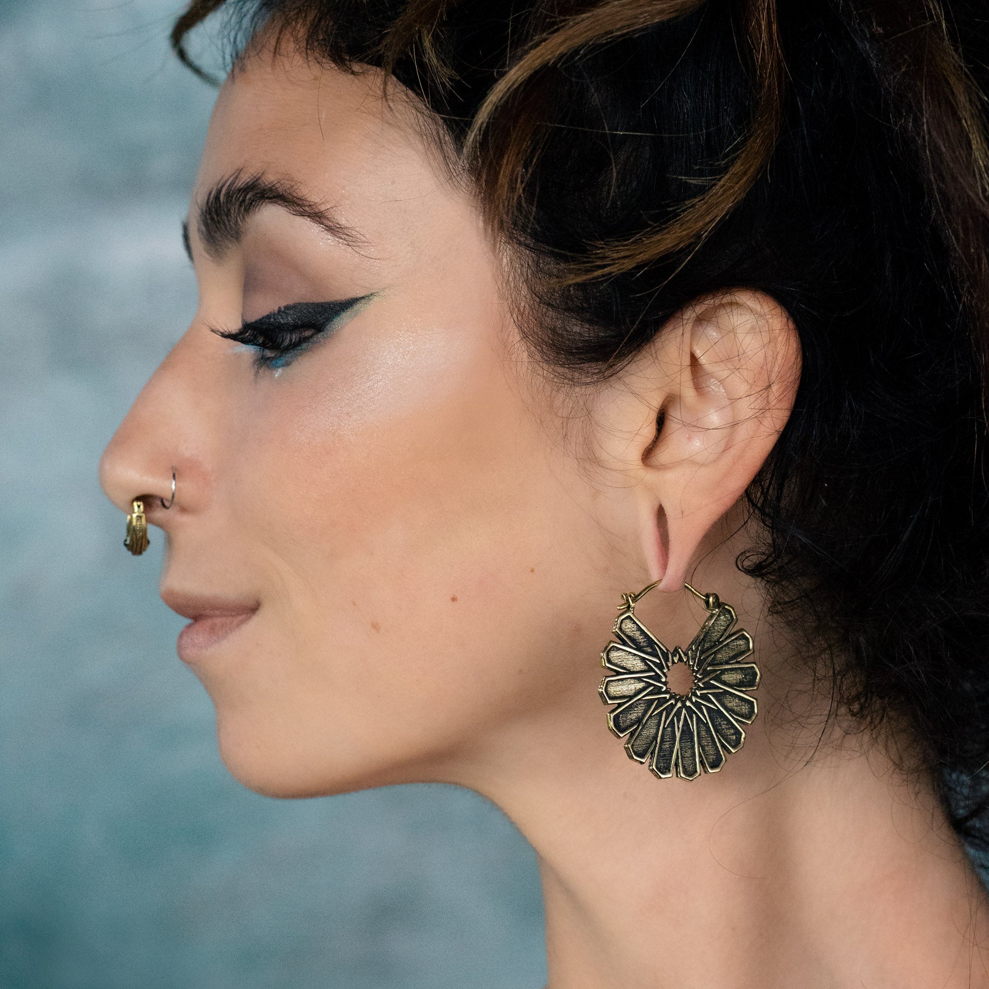 A person is shown in profile wearing the Kaya Movement MOSAIC Ethnic Mandala Disc Hoop Earrings in Gold. They have an intricate nose ring and dark, winged eyeliner. Their hair is tied up, and they are facing left against a blurred background.
