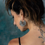 A person with wavy brown hair, pulled back, showcasing the MOSAIC Ethnic Mandala Disc Hoop Earrings in Silver (16 gauge) by Kaya Movement. They have a nose ring and a tattoo on the back of their neck and shoulder. Wearing a black strap top, they stand against a blurred blue-green background.