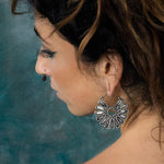 A woman with wavy brown hair is shown in profile against a blue-green background. She wears the large, ornate MOSAIC Ethnic Mandala Disc Hoop Earrings in Silver by Kaya Movement, evoking the essence of boho statement jewelry, along with a nose ring. Her makeup includes long, thick eyelashes and dark eyeliner.