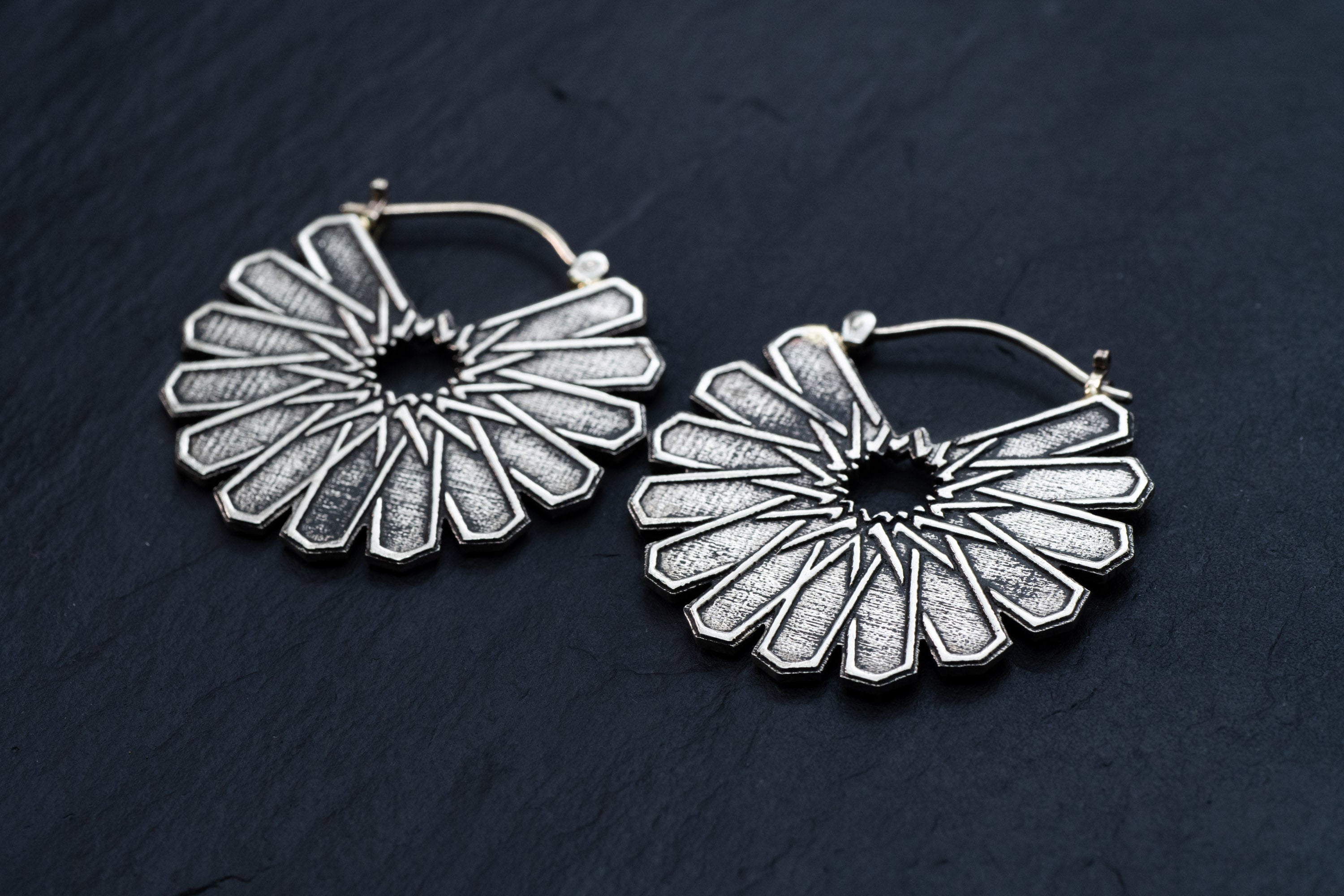 Close-up of a pair of MOSAIC Ethnic Mandala Disc Hoop Earrings in Silver by Kaya Movement, placed on a dark surface. These 16-gauge earrings feature a circular design with an intricate sunburst pattern, showcasing rays extending outward from a central point. The detailed texture captures and reflects light beautifully, highlighting the craftsmanship.