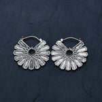 The MOSAIC Ethnic Mandala Disc Hoop Earrings in Silver by Kaya Movement lie on a dark, textured surface. These 16 gauge boho statement earrings feature an intricate geometric pattern with a central circular cutout and multiple triangular rays extending outward, creating a striking visual effect.