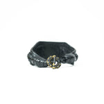 The ANOM Men's Black Cuff Leather Bracelet by Kaya Movement showcases textured, braided leather with an ornate golden clasp adorned with genuine Amethyst gemstone elements, secured at the front. Positioned against a plain white background, this piece embodies elegance and style.