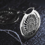 The CROP CIRCLE Oversized Tribal Disc Hoop Earrings in Silver by Kaya Movement feature an intricate circular design with geometric patterns and a sun-like motif at the center. One earring is showcased on a textured, dark surface, while a similar tribal earring can be seen blurred in the background.