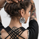 A person with tattoos on their arms and an intricate tattoo on the back of their neck is seen from behind. They are wearing Kaya Movement's CROP CIRCLE Oversized Tribal Disc Hoop Earrings in Silver, size 16 gauge, and a black outfit with crisscross straps on the back. Their hands are raised to adjust their hair.