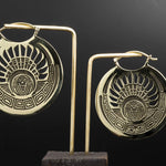 The CROP CIRCLE Oversized Tribal Disc Earrings in Gold by Kaya Movement are showcased against a black background. These golden, circular earrings feature an intricate sunburst pattern with a central face and geometric motifs along the edge. Each earring is displayed on a simple stand, highlighting their symmetrical design.