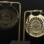 Two intricately designed Kaya Movement CROP CIRCLE Oversized Tribal Disc Earrings in Gold (16 gauge) are displayed against a black background. Each oversized gold hoop earring features a circular shape with an elaborate pattern that includes a central face, geometric lines, and sun-like rays extending outward. The earrings hang from a stand.