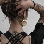A person with curly hair tied up in a bun is seen from the back, showcasing Kaya Movement's CROP CIRCLE Oversized Tribal Disc Earrings in Gold. They have tattoos on their arms and a tattoo on the upper back. The person is wearing a black top with crisscross straps.