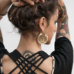 A person with a tattooed back and arms has their hair tied up and is placing their hands behind their head. They are wearing Kaya Movement's CROP CIRCLE Oversized Tribal Disc Earrings in Gold (16 gauge) and a black top with a lattice back design.