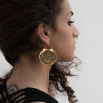 A person with curly hair and a side profile view, wearing large, Kaya Movement's CROP CIRCLE Oversized Tribal Disc Earrings in Gold, a septum piercing, and a silver ear cuff shaped like leaves. They have a tattoo visible on the back of their shoulder and are dressed in a black outfit against a plain wall.