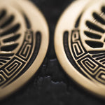 Close-up image of CROP CIRCLE Oversized Tribal Disc Earrings in Gold by Kaya Movement, showcasing intricate, symmetrical patterns with geometric shapes and curved, feather-like elements. The earrings are 16 gauge and set against a dark blurred background.