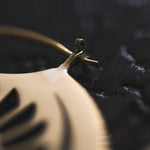 Close-up of the CROP CIRCLE Oversized Tribal Disc Earring in Gold by Kaya Movement, featuring a black feather-like design, resting on a textured black surface. The focus is on the detailed connection between the earring's hook and the main body, highlighting the intricate craftsmanship reminiscent of tribal statement earrings.