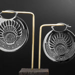 Two oversized, round earrings from Kaya Movement's "CROP CIRCLE" collection feature intricate, silver-colored tribal designs, embodying a boho style. These 16-gauge earrings showcase symmetrical patterns and a central face motif, elegantly hanging from thin gold rods against a dark, gradient background for contrast.
