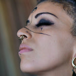 Close-up of a serene person with short hair, face in profile, eyes closed. They sport a septum ring from Kaya Movement's DARMA Fake Septum Ring or Ear Cuff in Gold, a nose stud, a lip ring, large hoop earrings, intricate black facial markings around the eyes and across the nose, and unisex nose jewelry that adds to their unique style.