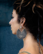 Profile view of a person with dark hair tied up in a messy bun, wearing the Circuit Oversized Visionary Disc Hoop Earrings in Silver (16 gauge) by Kaya Movement and a small nose ring. The person also has a tattoo on their back and is dressed in a light-colored tank top. The boho statement look is set against a blurred, dark blue background.