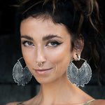 A woman with long brown hair tied up is wearing large, intricate Circuit Oversized Visionary Disc Hoop Earrings in Silver by Kaya Movement, a nose ring, and has striking green eyes. She is dressed casually with a confident expression and makeup accentuating her winged eyeliner. The background is dark and blurred.