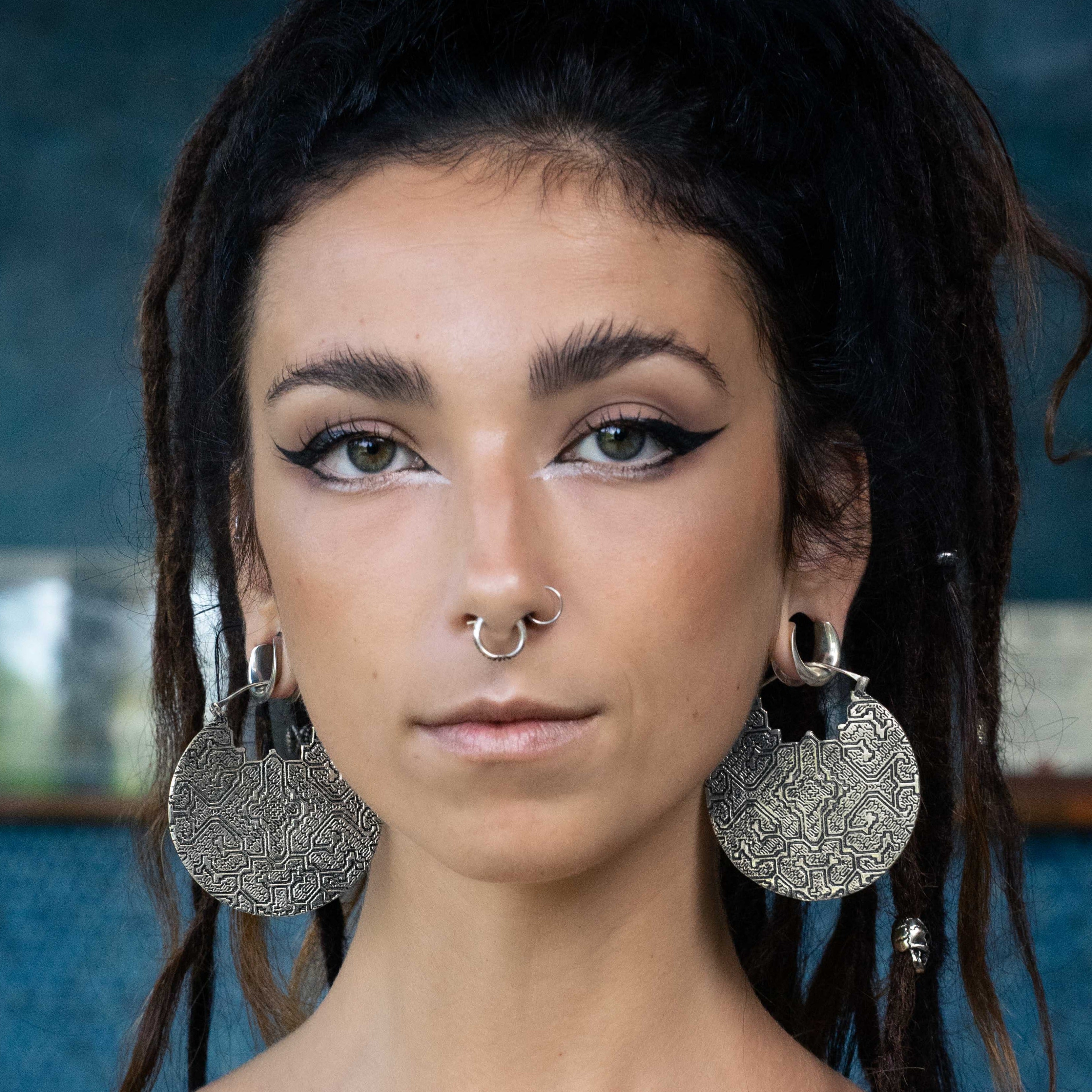 A person with long, dark hair styled in dreadlocks, wearing eyeliner with a dramatic wing, a septum piercing, and the PANCHA Oversized Tribal Shipibo Disc Earrings in Silver from Kaya Movement, looking directly at the camera with a neutral expression. The background is blurred and dark blue.