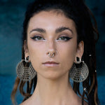 A person with dark, braided hair and a serious expression is seen against a blurred dark blue background. They wear Kaya Movement's ILLUSION Oversized Cyborg Sci Fi Disc Earrings in Silver, featuring intricate designs that emanate a futuristic aesthetic. Additionally, they sport a septum ring, two nose studs, and have bold, winged eyeliner.