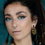 A person with medium skin tone, dark hair, and a nose piercing gazes confidently at the camera. They have bold eyeliner that extends past the eyes and turquoise eyeshadow. They wear TRILOBITE Large Biomechanical Clicker Ear Weights in Gold Brass by Kaya Movement and have dreadlocks. The background is teal.
