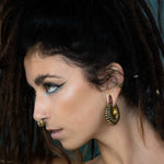 A woman with dark, curly hair styled in dreadlocks is shown in profile. She is wearing bold makeup with teal eyeliner and a dramatic wing. Her accessories include a biomechanical nose ring, the DARMA Fake Septum Ring or Ear Cuff in Gold by Kaya Movement, and spiral-shaped hoop earrings. The background is plain and dark.